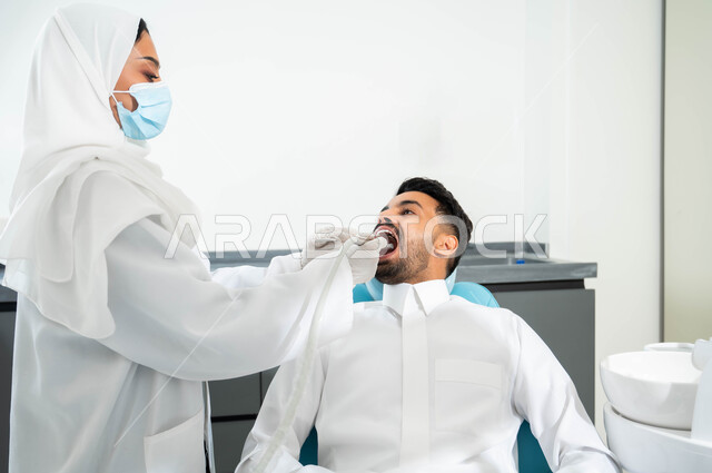 الادوات والمعدات اللازمة لإجراء الفحوصات الطبيه ,معالجة النخر والتسوس, شاب عربي خليجي سعودي يرتدي الزي التقليدي يجلس في العيادة السنية ويفتح فمه لتشخيص المرض, طبيبة عربية خليجيه سعودية ترتدي المعطف و القفازات والكمامة الطبية تقوم بفحص أسنان المريض