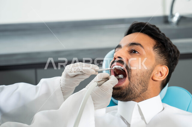 معالجة النخر والتسوس, صورة مقربة لشاب عربي خليجي سعودي يرتدي الزي التقليدي يجلس في العيادة السنية ويفتح فمه لتشخيص المرض, ايدي طبيبة عربية خليجيه سعودية ترتدي القفازات الطبية تقوم بفحص أسنان المريض, الادوات والمعدات اللازمة لإجراء الفحوصات الطبيه
