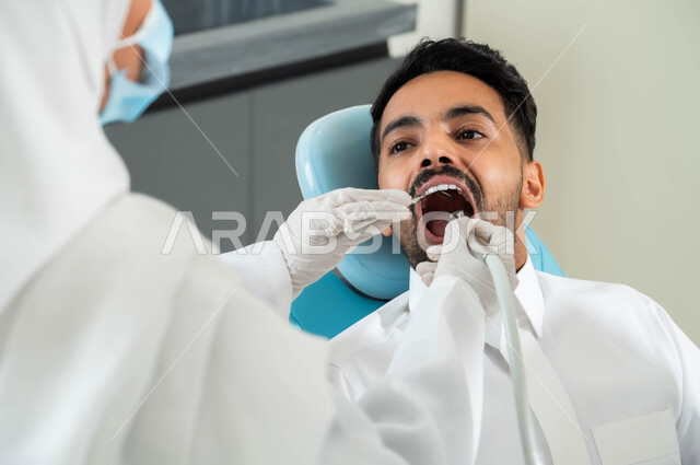 تركيب الحشوات و معالجة التسوس، جراحة الفم والأسنان، صورة مقربة من الخلف لطبيبة أسنان عربية خليجية سعودية تقوم بفحص أسنان المريض و تشخيص المرض, شاب عربي خليجي سعودي يرتدي الزي التقليدي و يجلس في العيادة السنية يقوم بإجراء الفحوصات الدورية للعناية بالصحة الفموية
