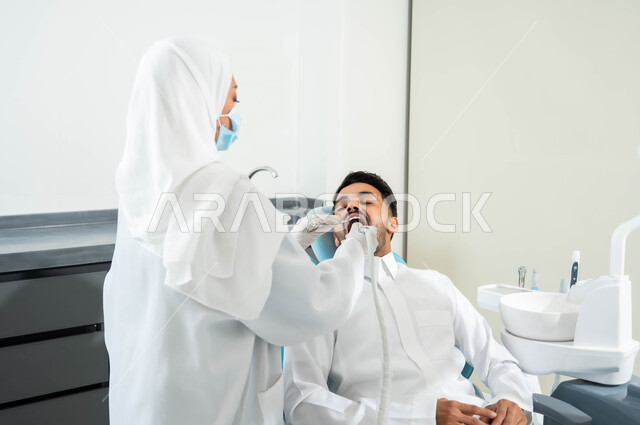 معالجة التسوس وتركيب الحشوات، المعدات والأدوات اللازمة لجراحة الفم، صورة مقربة من الخلف لطبيبة عربية خليجية سعودية تقوم بفحص أسنان المريض و تشخيص المرض، شاب عربي خليجي سعودي يرتدي الزي التقليدي يجلس في العيادة السنية يقوم بإجراء الفحوصات للعناية بالصحة الفموية