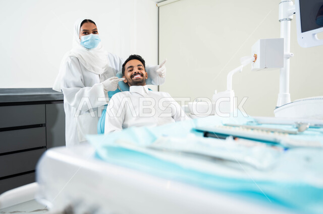 النظر الى الكاميرا بايماءات السرور ,استخدام قشور فينير من الخزف ,الاهتمام في الصحة الفموية ,طبيبة أسنان عربية خليجية سعودية ترتدي المعطف و القفازات والكمامة الطبية تشير بيدها الى إبتسامة هوليود لدى شاب عربي خليجي سعودي يرتدي الزي التقليدي يجلس في العيادة السنية