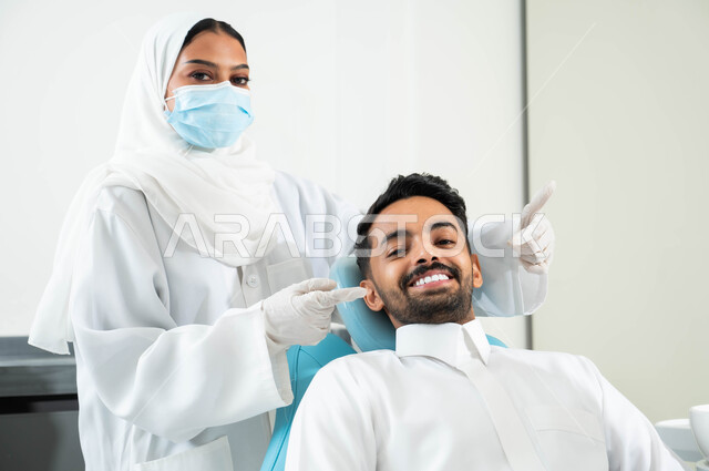 استخدام قشور فينير من الخزف ,الاهتمام في الصحة الفموية ,صورة مقربة لطبيبة أسنان عربية خليجية سعودية ترتدي القفازات والكمامة الطبية تشير بيدها الى إبتسامة هوليود لدى شاب عربي خليجي سعودي يرتدي الزي التقليدي يجلس في العيادة السنية, النظر الى الكاميرا بايماءات السرور