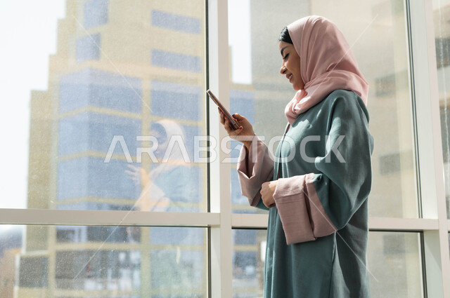 إستخدام اجهزة تقنية حديثة, تصفح وسائل التواصل الاجتماعي باستخدام الجوال, صورة مقربة من الجانب لإمرأة عربية سعودية خليجية محجبة تقف بجانب نوافذ زجاجية كبيرة في إحدى الأبراج الراقية، تدوين البيانات والملاحظات على الهاتف المحمول, التطبيقات التقنية للأجهزة اللوحية