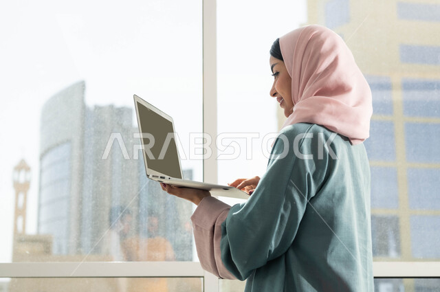 احتراف وظائف الحاسوب وتكنلوجيا المعلومات, صورة مقربة من الجانب لإمرأة عربية سعودية خليجية محجبة تقف بجانب نوافذ زجاجية كبيرة في إحدى الأبراج الراقية تحمل بيدها اللابتوب, إنجاز مهام العمل, التطبيقات التقنية للأجهزة اللوحية, مفهوم إدارة المشاريع عن بعد