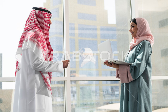 مهن ووظائف سعودية, ضمان فهم واضح وشامل للاعمال, الحوارات والنقاشات ضمن الفريق, مفهوم التعاون في العمل, صورة لرجل وامرأة عربيان سعوديان خليجيان يرتديان الزي التقليدي السعودي بالنقاش في الشركة الراقية بجانب النافذة الزجاجية, استخدام الاجهزة والتقنيات المتطورة