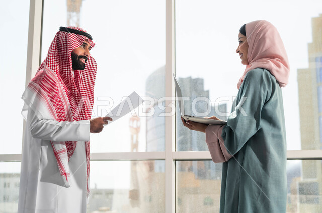 Using advanced equipment and technologies, ensuring a clear and comprehensive understanding of the business, dialogues and discussions within the team, the concept of cooperation at work, a picture of a Saudi Gulf Arab man and woman wearing traditional Saudi dress, discussing in the upscale company next to the glass window, Saudi professions and jobs.