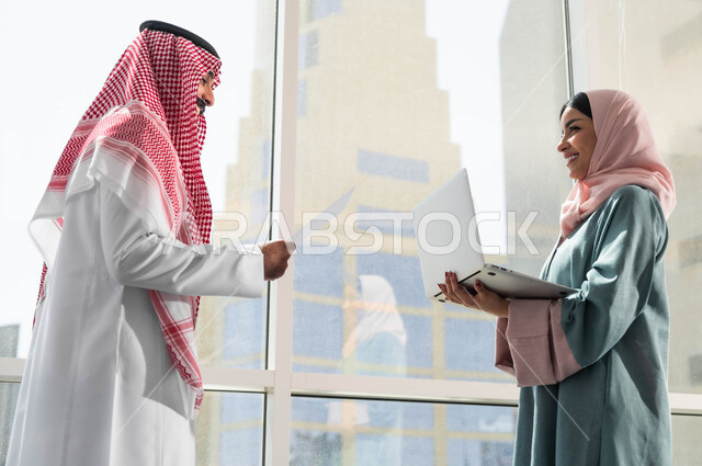 The concept of cooperation at work, ensuring a clear and comprehensive understanding of the business, dialogues and discussions within the team, a picture of a Saudi Gulf Arab man and woman wearing traditional Saudi dress, discussing in the upscale company next to the glass window, using advanced devices and technologies, Saudi professions and jobs