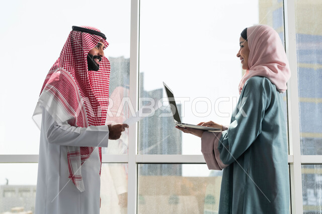 الحوارات والنقاشات ضمن الفريق, مفهوم التعاون في العمل, صورة لرجل وامرأة عربيان سعوديان خليجيان يرتديان الزي التقليدي السعودي بالنقاش في الشركة الراقية بجانب النافذة الزجاجية, استخدام الاجهزة والتقنيات المتطورة, مهن ووظائف سعودية, ضمان فهم واضح وشامل للاعمال