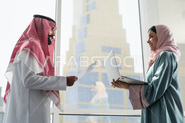ضمان فهم واضح وشامل للاعمال, الحوارات والنقاشات ضمن الفريق, مفهوم التعاون في العمل, صورة لرجل وامرأة عربيان سعوديان خليجيان يرتديان الزي التقليدي السعودي بالنقاش في الشركة الراقية بجانب النافذة الزجاجية, استخدام الاجهزة والتقنيات المتطورة, مهن ووظائف سعودية