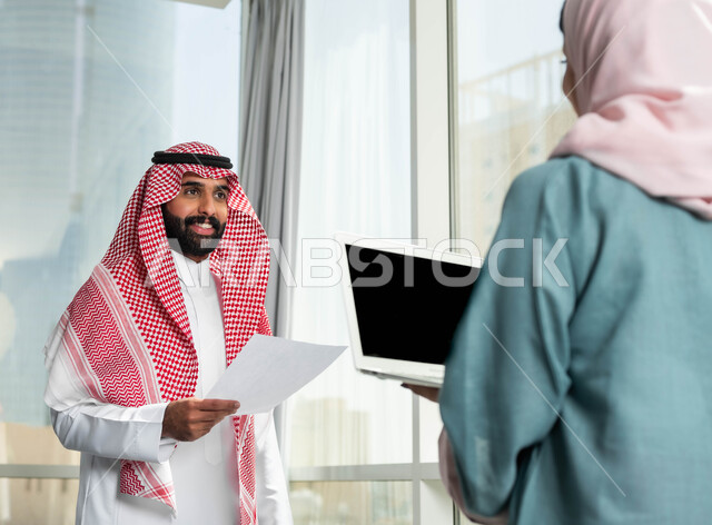 تحقيق استراتيجيات واهداف العمل, مناقشة ومتابعة الاعمال, صورة لرجل وامرأة عربيان سعوديان خليجيان يرتديان الزي التقليدي يقومان بالحوارات في الشركة الراقية بجانب النافذة الزجاجية, استخدام الاجهزة والتقنيات الحديثة المتطورة, التعاون والتفاهم بين الزملاء