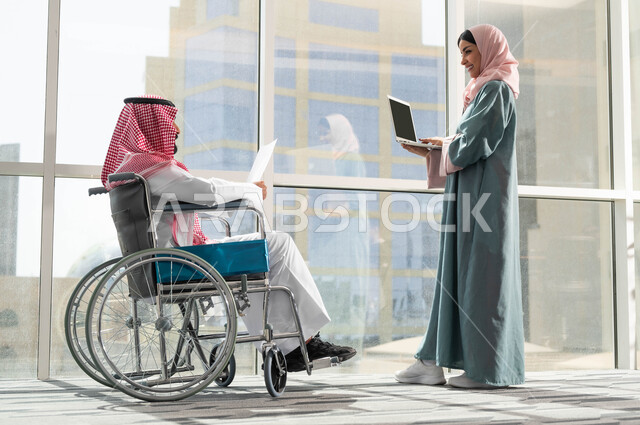 استخدام الاجهزة والتقنيات الحديثة, صورة من الجانب لرجل وامرأة عربيان سعوديان خليجيان يرتديان الزي التقليدي السعودي يقومان بالحوارات في الشركة الراقية بجانب النافذة الزجاجية, مناقشة ومتابعة الاعمال وتحقيق الاهداف المطلوبة, توفير المستلزمات الخاصة بالمقعدين
