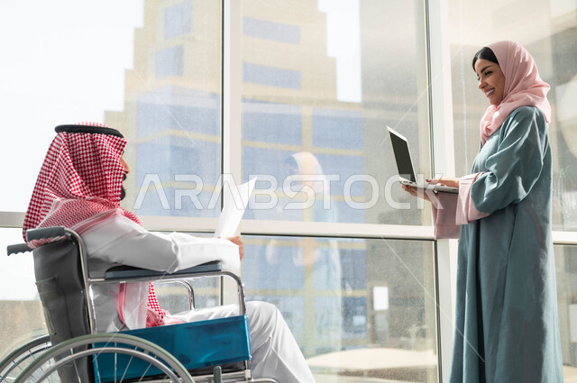 مناقشة ومتابعة الاعمال وتحقيق الاهداف المطلوبة, توفير المستلزمات الخاصة بالمقعدين, استخدام الاجهزة والتقنيات الحديثة, صورة من الجانب لرجل وامرأة عربيان سعوديان خليجيان يرتديان الزي التقليدي السعودي يقومان بالحوارات في الشركة الراقية بجانب النافذة الزجاجية