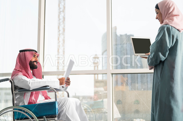 توفير المستلزمات الخاصة بالمقعدين, استخدام الاجهزة والتقنيات الحديثة, صورة من الجانب لرجل وامرأة عربيان سعوديان خليجيان يرتديان الزي التقليدي السعودي يقومان بالحوارات في الشركة الراقية بجانب النافذة الزجاجية, مناقشة ومتابعة الاعمال وتحقيق الاهداف المطلوبة