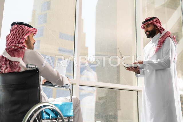 تأمين اجهزة ومعدات تساعد بالحركة والتنقل, النجاح في تحقيق أهداف واستراتيجيات الشركة, صورة من الجانب لموظفان عربيان خليجيان سعوديان يرتديان الزي التقليدي السعودي يقومان بالحوارات والنقاشات في الشركة الراقية بجانب النافذة الزجاجية, مفهوم التركيز والدقة في الإنجاز