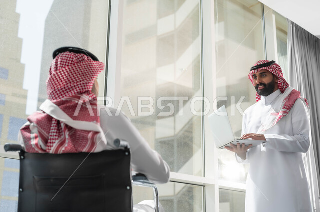 استخدام الحاسب المحمول والتكنولوجيا، نمط الحياة في الشركات السعودية، اتاحة المجال امام ذوي الهمم للدخول الى مجال العمل، صورة مقربة لرجلين عربيين خليجيين سعوديين يرتديان الشماغ والثوب التقليدي يقومان بتبادل ومشاركة المعلومات والبيانات داخل مقر الشركة، التعاون والمودة بين الموظفين
