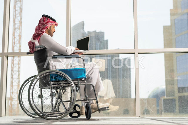 دمج وتوظيف ذوي الهمم في مختلف المجالات العملية, رجل عربي سعودي خليجي مبتسم يرتدي الشماغ والثوب التقليدي يجلس على الكرسي المتحرك يعمل على الحاسب،  استخدام الأجهزة التقنية والتكنولوجية في مقر العمل، دعم واهتمام المجتمع السعودي بأصحاب الاحتياجات الخاصه