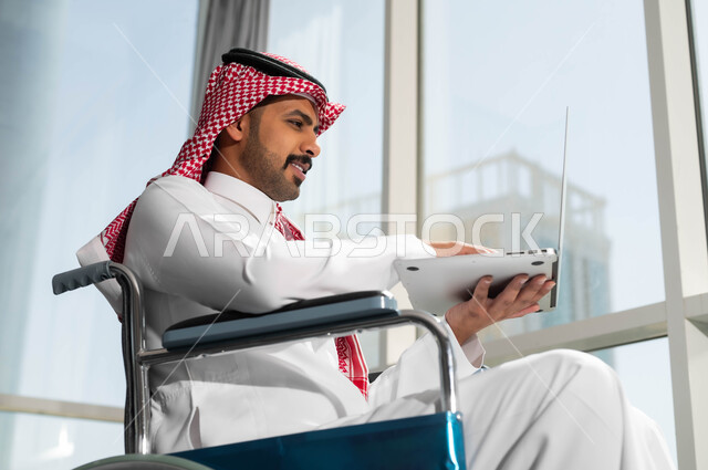دمج وتوظيف ذوي الهمم في مختلف المجالات العملية, صورة مقربة لرجل عربي سعودي خليجي مبتسم يرتدي الشماغ والثوب التقليدي يجلس على الكرسي المتحرك يعمل على الحاسب،  استخدام الأجهزة التقنية والتكنولوجية في مقر العمل، دعم واهتمام المجتمع السعودي بأصحاب الاحتياجات الخاصه