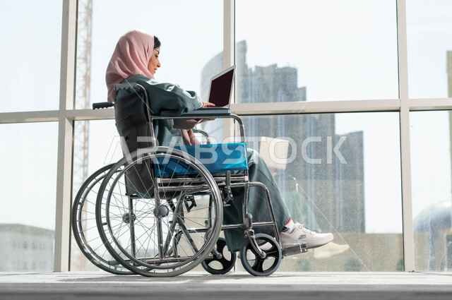 دعم واهتمام المجتمع السعودي بأصحاب الاحتياجات الخاصه, دمج وتوظيف ذوي الهمم في مختلف المجالات العملية, صورة من الجانب لإمرأة عربية سعودية خليجية مبتسمة ترتدي الحجاب تجلس على الكرسي المتحرك تمسك الحاسوب المحمول، استخدام الأجهزة التقنية والتكنولوجية في مقر العمل