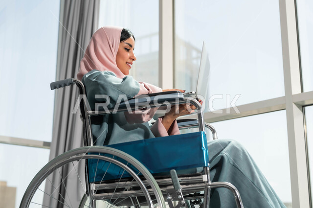 دمج وتوظيف ذوي الهمم في مختلف المجالات العملية, صورة مقربة لإمرأة عربية سعودية خليجية مبتسمة ترتدي الحجاب تجلس على الكرسي المتحرك تمسك الحاسوب المحمول، استخدام الأجهزة التقنية والتكنولوجية في مقر العمل، دعم واهتمام المجتمع السعودي بأصحاب الاحتياجات الخاصه