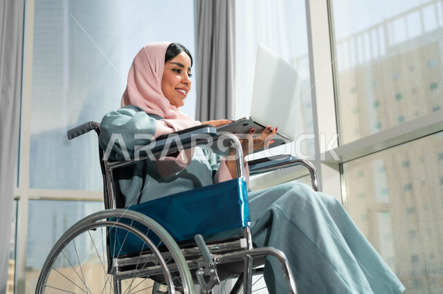 استخدام الأجهزة التقنية والتكنولوجية في مقر العمل، دعم واهتمام المجتمع السعودي بأصحاب الاحتياجات الخاصه، دمج وتوظيف ذوي الهمم في مختلف المجالات العملية, صورة مقربة لإمرأة عربية سعودية خليجية مبتسمة ترتدي الحجاب تجلس على الكرسي المتحرك تمسك الحاسوب المحمول