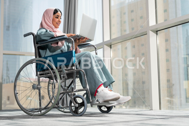 Saudi society’s interest and support for people with special needs, integrating and employing people with special needs in various practical fields, a smiling Saudi Arabian Gulf woman wearing the hijab sitting in a wheelchair holding a laptop, using technical and technological devices in the workplace