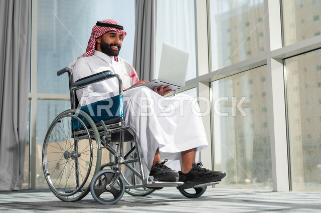 دمج وتوظيف ذوي الهمم في مختلف المجالات العملية, رجل عربي سعودي خليجي مبتسم يرتدي الشماغ والثوب التقليدي يجلس على الكرسي المتحرك يعمل على الحاسب،  استخدام الأجهزة التقنية والتكنولوجية في مقر العمل، دعم واهتمام المجتمع السعودي بأصحاب الاحتياجات الخاصه