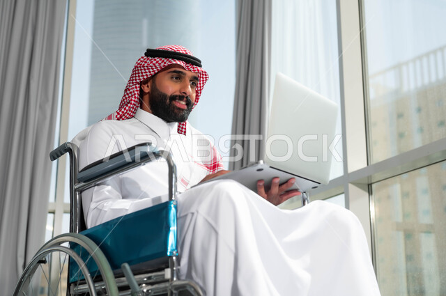 استخدام الأجهزة التقنية والتكنولوجية في مقر العمل، دعم واهتمام المجتمع السعودي بأصحاب الاحتياجات الخاصه، دمج وتوظيف ذوي الهمم في مختلف المجالات العملية, صورة مقربة لرجل عربي سعودي خليجي مبتسم يرتدي الشماغ والثوب التقليدي يجلس على الكرسي المتحرك يعمل على الحاسب