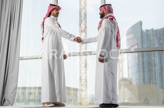 Concluding agreements and business deals, getting to know each other and building social relationships, welcoming and receiving gestures of happiness and pleasure, two smiling Saudi Gulf Arab men wearing the traditional Saudi shemagh and thobe, shaking hands with each other, expressions of pleasure in reaching the goals.