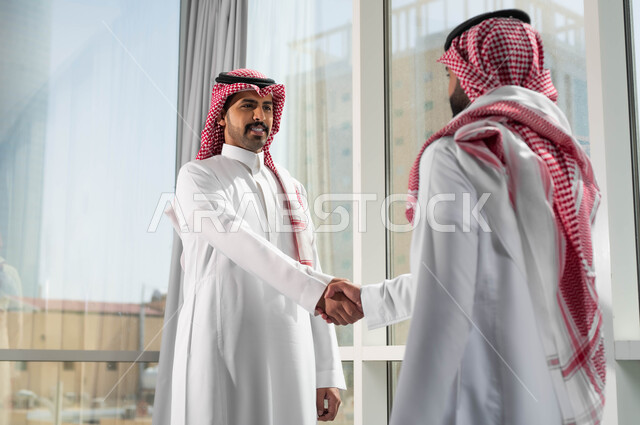 التعارف وبناء العلاقات الاجتماعية، الترحيب والاستقبال بإيماءات السعادة والسرور، صورة مقربة لرجلين عربيين سعوديين خليجين مبتسمين يرتديان الشماغ والثوب السعودي التقليدي يقومان بمصافحة بعضهما، تعابير السرور بالوصول الى الأهداف، عقد الاتفاقات والصفقات التجارية