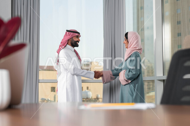 مفهوم المساواة بين النساء والرجال، رجل عربي خليجي سعودي  يرتدي الشماغ والثوب التقليدي يقوم بمصافحة إمرأة ترتدي الحجاب والعباءة في غرفة الاجتماع داخل مقر الشركة، الترحيب والاستقبال بالموظفين الجدد، بناء علاقات ايجابية مع زملاء العمل، اجراء مقابلة التعيين والتوظيف