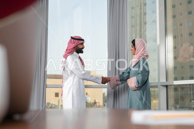 الترحيب والاستقبال بالموظفين الجدد، بناء علاقات ايجابية مع زملاء العمل، اجراء مقابلة التعيين والتوظيف، مفهوم المساواة بين النساء والرجال، رجل عربي خليجي سعودي يرتدي الشماغ والثوب التقليدي يقوم بمصافحة إمرأة ترتدي الحجاب والعباءة في غرفة الاجتماع داخل مقر الشركة