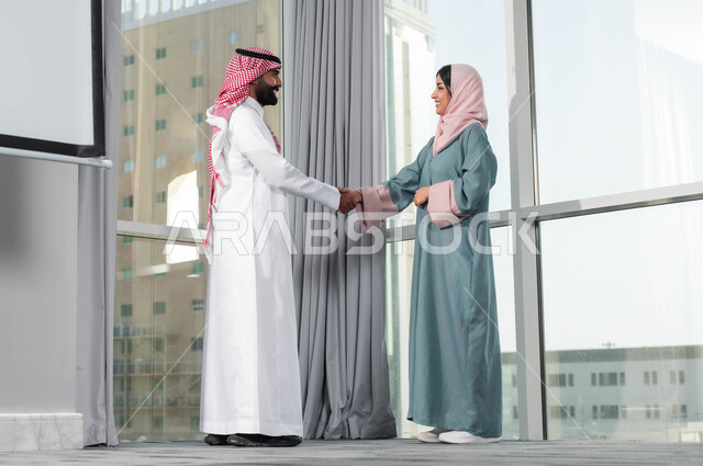 بناء علاقات ايجابية مع زملاء العمل، اجراء مقابلة التعيين والتوظيف، مفهوم المساواة بين النساء والرجال، رجل عربي خليجي سعودي يرتدي الشماغ والثوب التقليدي يقوم بمصافحة إمرأة ترتدي الحجاب والعباءة في غرفة الاجتماع داخل مقر الشركة، الترحيب والاستقبال بالموظفين الجدد