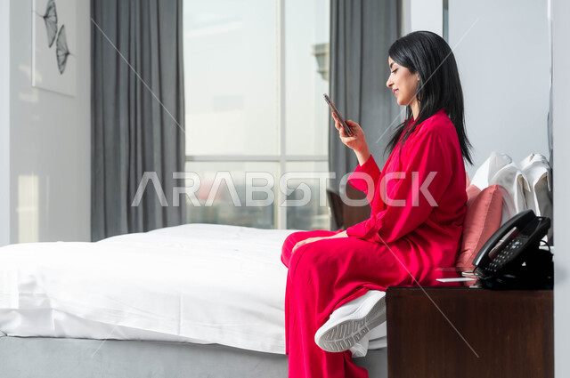 Expressions and gestures of happiness and pleasure using advanced technical devices, staying in one of the Kingdom’s luxury hotels, a side photo of a Saudi Gulf Arab woman sitting on a bed in the bedroom using a mobile phone.