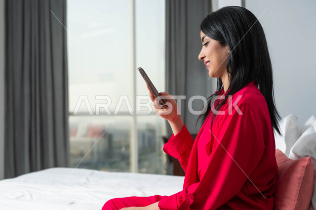 الإقامة في إحدى فنادق المملكة الفاخرة، ايماءات وتعابير السعادة و السرور باستخدام الأجهزة التقنية المتطورة، صورة من الجانب لامرأة عربية خليجية سعودية تجلس على سرير في غرفة النوم تستخدم الهاتف المحمول