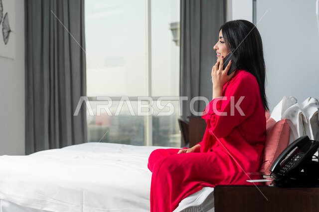 تبادل الاخبار والأحاديث، محادثات ودردشات مع الاصدقاء، الإقامة في إحدى فنادق المملكة الفاخرة، ايماءات السعادة و السرور بإجراء مكالمة هاتفية، صورة من الجانب لامرأة عربية خليجية سعودية تجلس على سرير في غرفة النوم تقوم بالتحدث عبر الهاتف المحمول