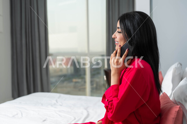 محادثات ودردشات مع الاصدقاء، الإقامة في إحدى فنادق المملكة الفاخرة، ايماءات السعادة و السرور بإجراء مكالمة هاتفية، صورة من الجانب لامرأة عربية خليجية سعودية تجلس على سرير في غرفة النوم تقوم بالتحدث عبر الهاتف المحمول، قضاء اوقات ممتعة بتبادل الاخبار والأحاديث
