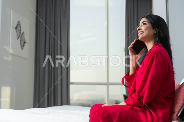 محادثات ودردشات مع الاصدقاء، الإقامة في إحدى فنادق المملكة الفاخرة، ايماءات السعادة و السرور بإجراء مكالمة هاتفية، صورة من الجانب لامرأة عربية خليجية سعودية تجلس على سرير في غرفة النوم تقوم بالتحدث عبر الهاتف المحمول، قضاء اوقات ممتعة بتبادل الاخبار والأحاديث