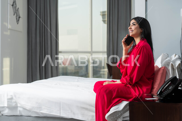 الإقامة في إحدى فنادق المملكة الفاخرة، ايماءات السعادة و السرور بإجراء مكالمة هاتفية، صورة من الجانب لامرأة عربية خليجية سعودية تجلس على سرير في غرفة النوم تقوم بالتحدث عبر الهاتف المحمول، قضاء اوقات ممتعة بتبادل الاخبار والأحاديث، محادثات ودردشات مع الاصدقاء