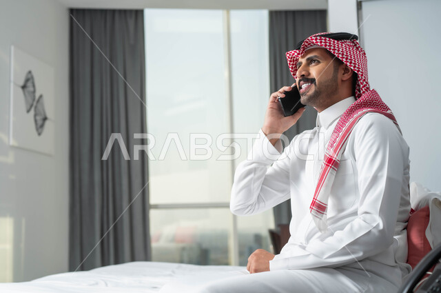 تبادل الاحاديث المسلية والحوارات، استخدام الأجهزة التكنولوجية الحديثة والمتطورة، رجل عربي خليجي سعودي مبتسم يرتدي الشماغ والثوب التقليدي يجلس على السرير في إحدى الفنادق الراقية يقوم بإجراء مكالمة صوتيه عبر الهاتف المحمول