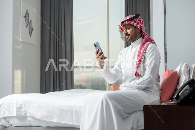 قضاء الإجازة الصيفية في إحدى الفنادق الراقية، استخدام الأجهزة التكنولوجية الحديثة والمتطورة، رجل عربي خليجي سعودي يرتدي الشماغ والثوب التقليدي يجلس على السرير في غرفة النوم ويستخدم الهاتف المحمول بإيماءات الاندماج والتركيز