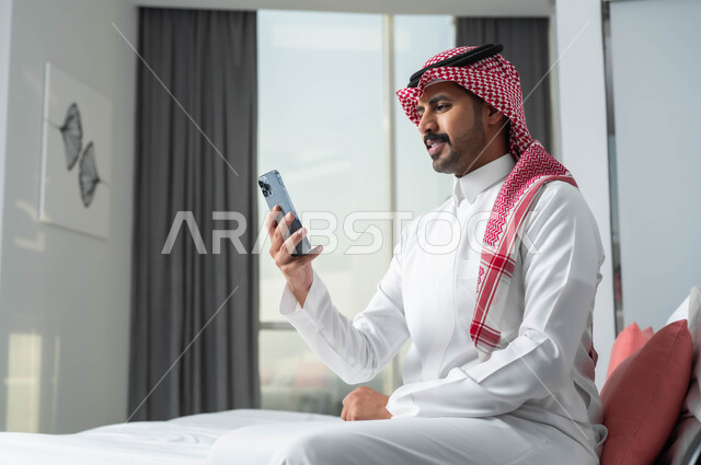 قضاء الإجازة الصيفية في إحدى الفنادق الراقية، استخدام الأجهزة التكنولوجية الحديثة والمتطورة، رجل عربي خليجي سعودي يرتدي الشماغ والثوب التقليدي يجلس على السرير في غرفة النوم ويستخدم الهاتف المحمول بإيماءات الاندماج والتركيز