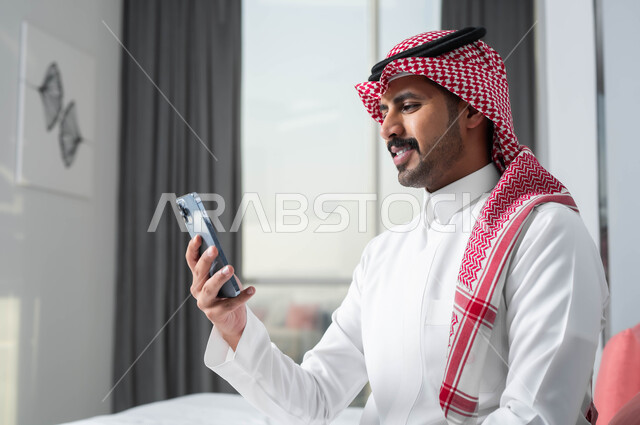 تعابير وإيماءات الاندماج والتركيز، قضاء الإجازة الصيفية في إحدى الفنادق الراقية، استخدام الأجهزة التكنولوجية الحديثة والمتطورة، صورة مقربة لرجل عربي خليجي سعودي يرتدي الشماغ والثوب التقليدي يجلس على السرير في غرفة النوم ويستخدم الهاتف المحمول