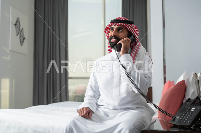 التواصل مع أحد أقسام الفندق، الدردشة وتبادل الاحاديث المسلية، استخدام الأجهزة السلكية للتواصل، رجل عربي خليجي سعودي مبتسم يرتدي الشماغ والثوب التقليدي يجلس على السرير في إحدى الفنادق الراقية يقوم بإجراء اتصال عبر الهاتف الثابت