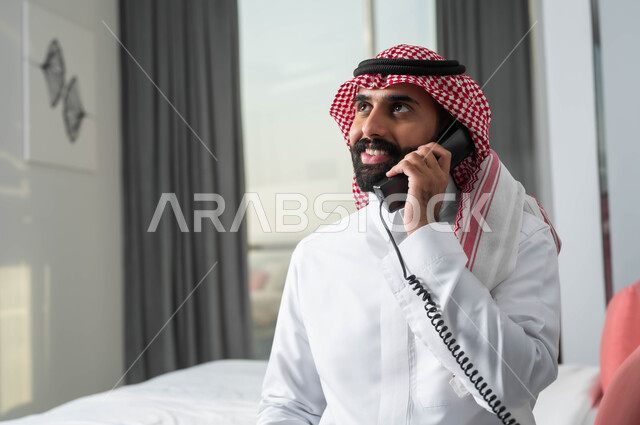 الدردشة وتبادل الاحاديث المسلية، استخدام الأجهزة السلكية للتواصل، صورة مقربة لرجل عربي خليجي سعودي مبتسم يرتدي الشماغ والثوب التقليدي يجلس على السرير في إحدى الفنادق الراقية يقوم بإجراء اتصال عبر الهاتف الثابت، التواصل مع أحد أقسام الفندق