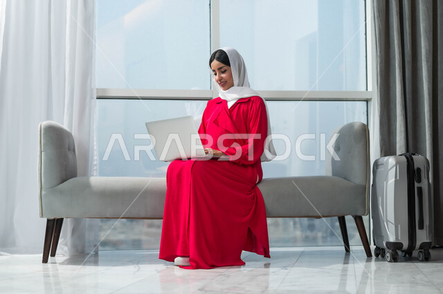 جولة عمل خارجية، استخدام الأجهزة التكنولوجية الحديثة والمتطورة، إمرأة عربية خليجية سعودية مبتسمة ترتدي الحجاب تجلس في لوبي فندق فاخر بجانب حقيبة السفر، تعابير وايماءات الاندماج والتركيز لإنجاز المهام، إجراء الحجوزات الفندقية وتذاكر الطيران اونلاين عبر اللابتوب