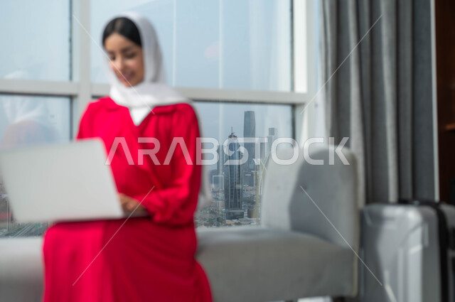 جولة عمل خارجية، استخدام الأجهزة التكنولوجية الحديثة والمتطورة، إمرأة عربية خليجية سعودية مبتسمة ترتدي الحجاب تجلس في لوبي فندق فاخر بجانب حقيبة السفر، تعابير وايماءات الاندماج والتركيز لإنجاز المهام، إجراء الحجوزات الفندقية وتذاكر الطيران اونلاين عبر اللابتوب