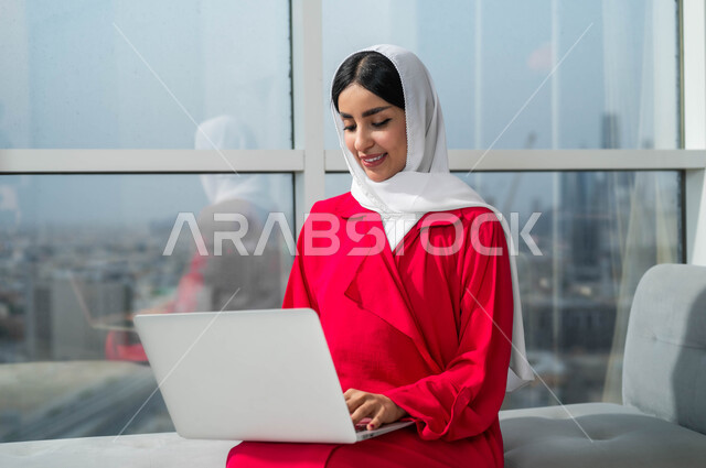 تعابير وايماءات الاندماج والتركيز لإنجاز المهام والأعمال، جولة عمل خارجية، استخدام الأجهزة التكنولوجية الحديثة والمتطورة، صورة مقربة لإمرأة عربية خليجية سعودية مبتسمة ترتدي الحجاب تجلس في لوبي فندق فاخر تقوم بالعمل على الحاسوب المحمول