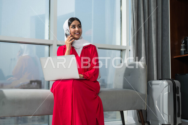 إجراء الحجوزات الفندقية وتذاكر الطيران اونلاين عبر اللابتوب، جولة عمل خارجية، استخدام الأجهزة التكنولوجية الحديثة والمتطورة، إمرأة عربية خليجية سعودية مبتسمة ترتدي الحجاب تجلس في لوبي فندق فاخر بجانب حقيبة السفر تنظر الى شيء ما، الرد على اتصالات الهاتف المحمول
