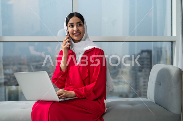 Using modern and advanced technological devices, a close-up picture of a smiling Saudi Arabian Gulf woman wearing a hijab, working on a laptop, chatting and talking on a mobile phone, looking at something while making a phone call on a mobile phone, sitting in the lobby of an upscale hotel.