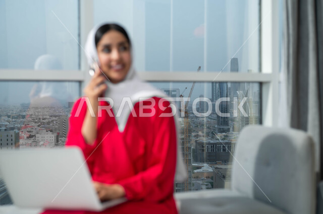 الجلوس في لوبي إحدى الفنادق الراقية، استخدام الأجهزة التكنولوجية الحديثة والمتطورة، صورة مقربة لإمرأة عربية خليجية سعودية مبتسمة ترتدي الحجاب تعمل على اللابتوب وتقوم بالدردشة والتحدث على الهاتف المحمول، النظر إلى شيء ما أثناء إجراء مكالمة هاتفية عبر الجوال